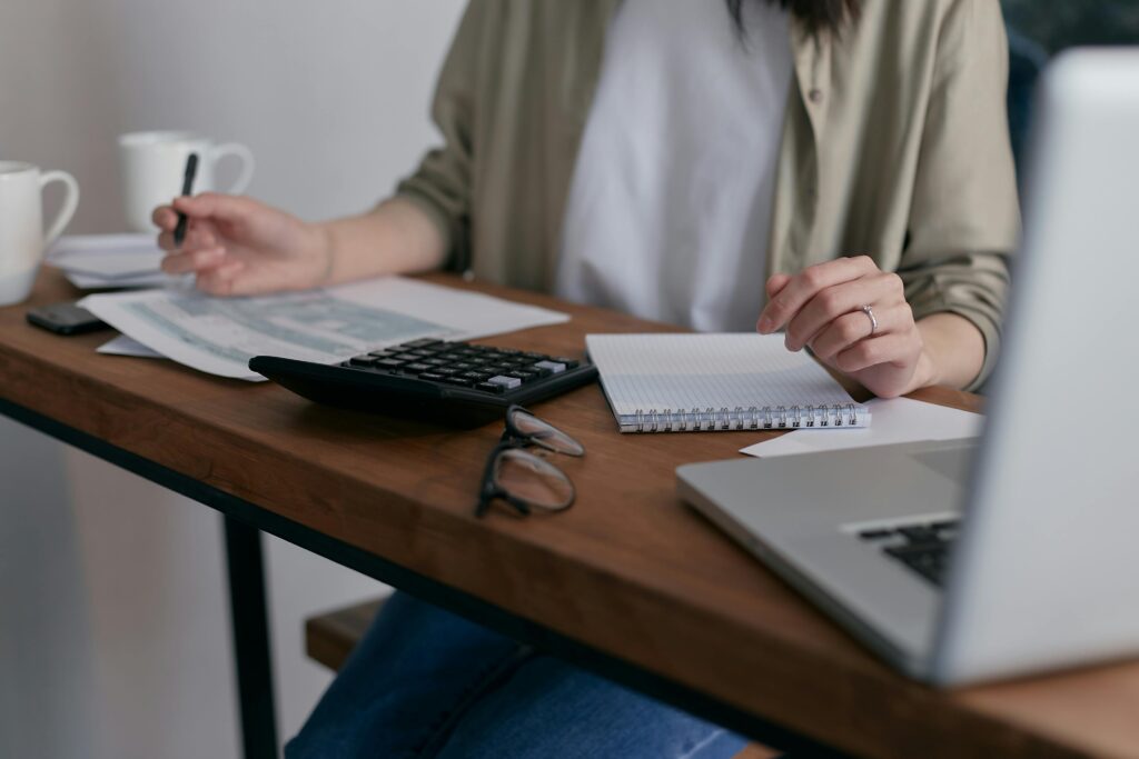 pexels-photo-6963857-6963857 A woman manages finances at home, using a laptop and calculator on a wooden desk.